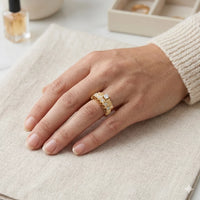 Hand wearing a gold ring with a textured band on a light fabric surface.